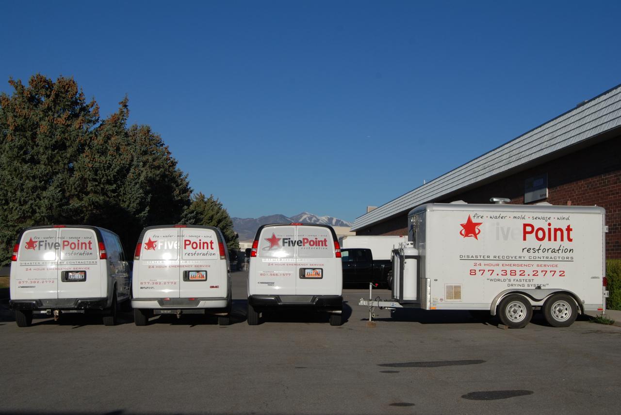 Three branded Five Point Restoration white GMC vans and equipment trailer lined up in a parking lot with Utah mountains in the background